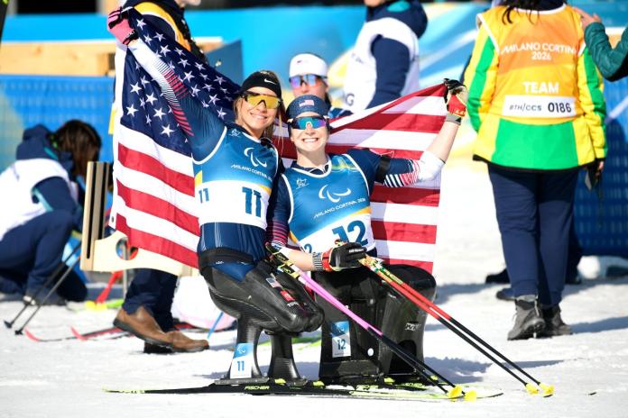 Two female sit-skiers pose for a photo by holding the flag of the USA
